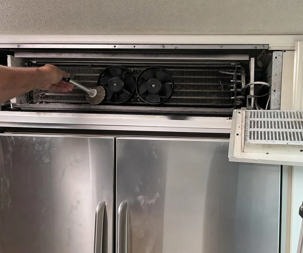A hand brushing dust from the fins of a refrigerator condenser coil behind its opened grille