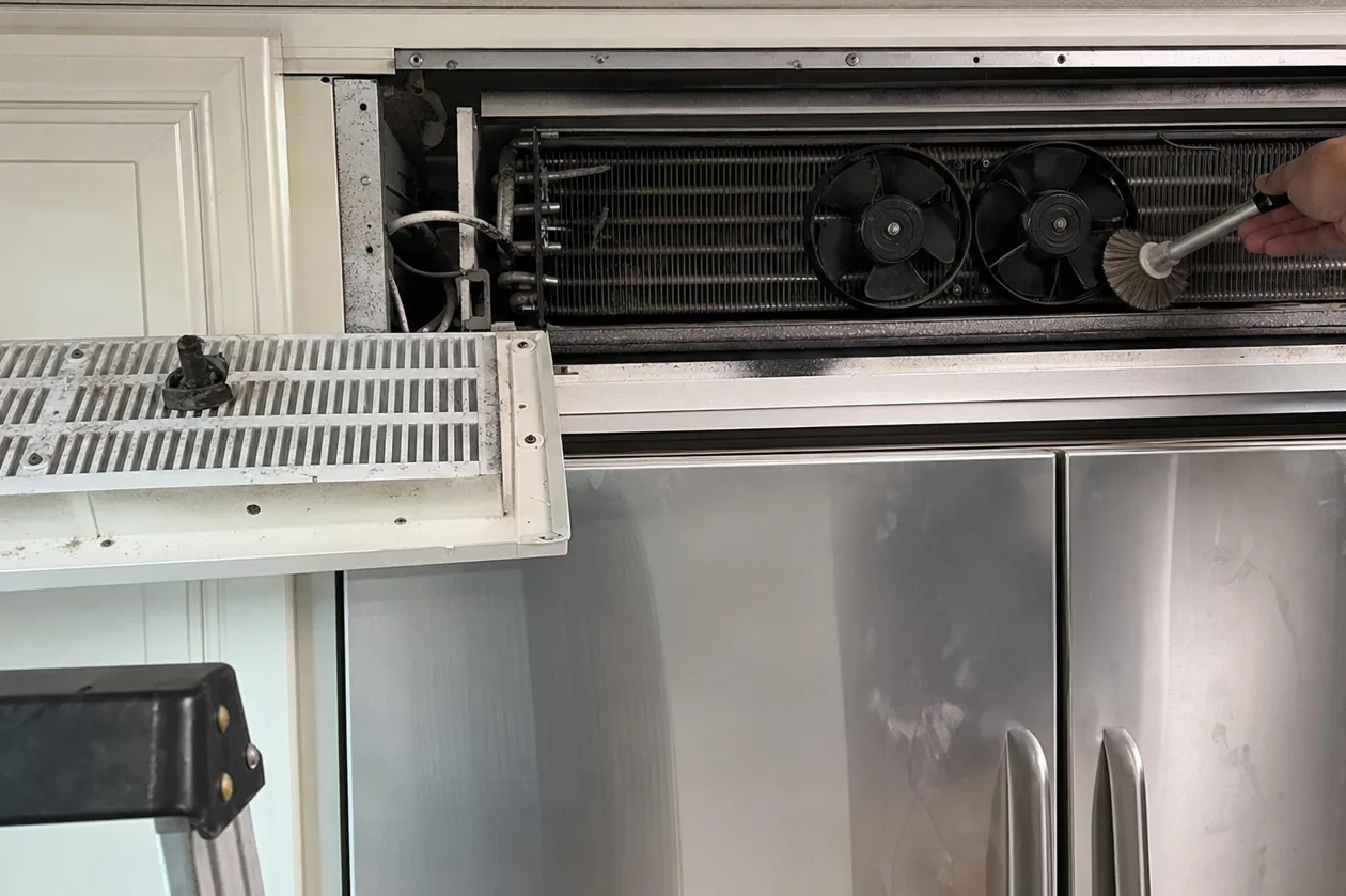 The exposed condensing unit above a stainless refrigerator, its grille panel hinged open for cleaning